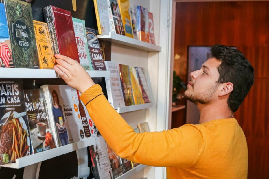 Autor Naican Escobar, com blusa amarela, em frente a uma estante com muitos livros
